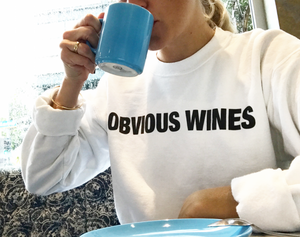 Person sipping coffee wearing white sweatshirt with the text Obvious Wines on the front